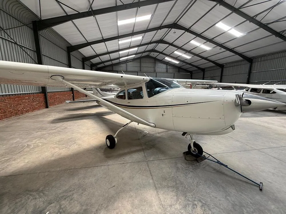 A white Cessna Skylane aircraft with blue and gold stripes parked in a hangar with other aircraft in the background.
