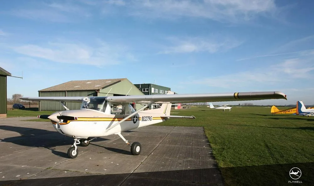 A front-right view of a white and beige Cessna 172 with yellow and black stripes, parked on a concrete pad at an airfield.