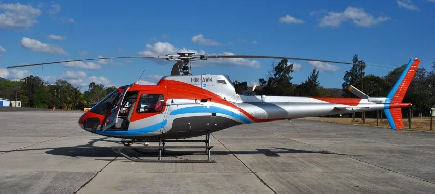 A red, silver, and blue helicopter with registration HR-AWK is parked on an airfield under a clear blue sky.