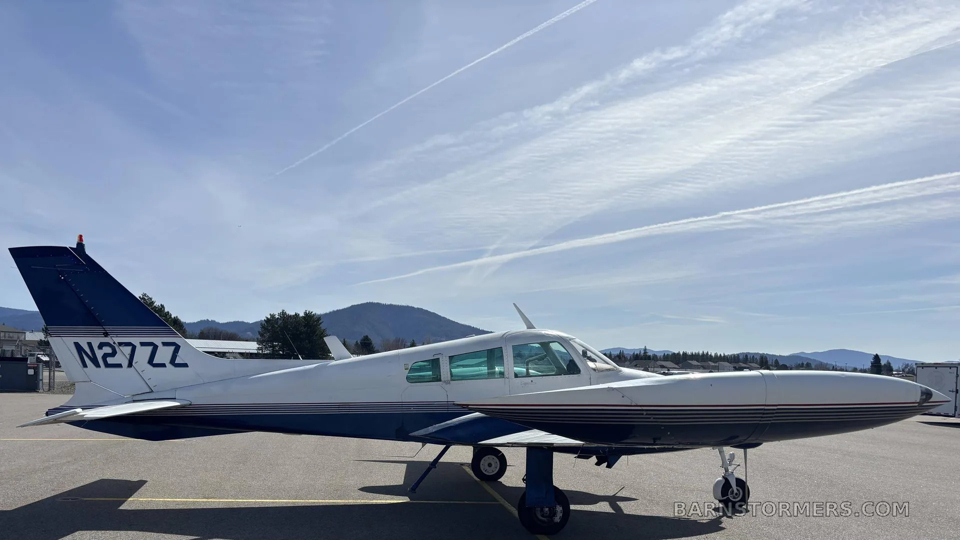 A white and blue Cessna 310I, registration N27ZZ, parked on an airfield with mountains and contrails in the sky.