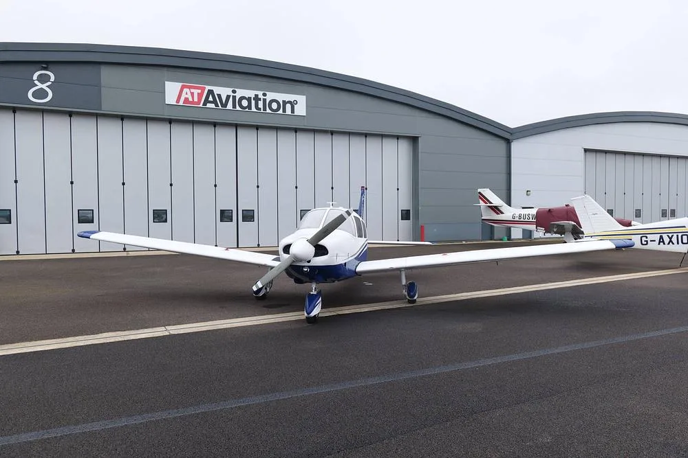 A white and blue Piper Cherokee aircraft parked on an airfield in front of an AT Aviation hangar, with another aircraft in the background.