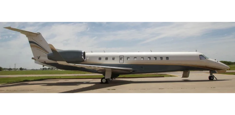 A white and dark grey business jet with gold accent stripes is parked on a tarmac under a partly cloudy sky.