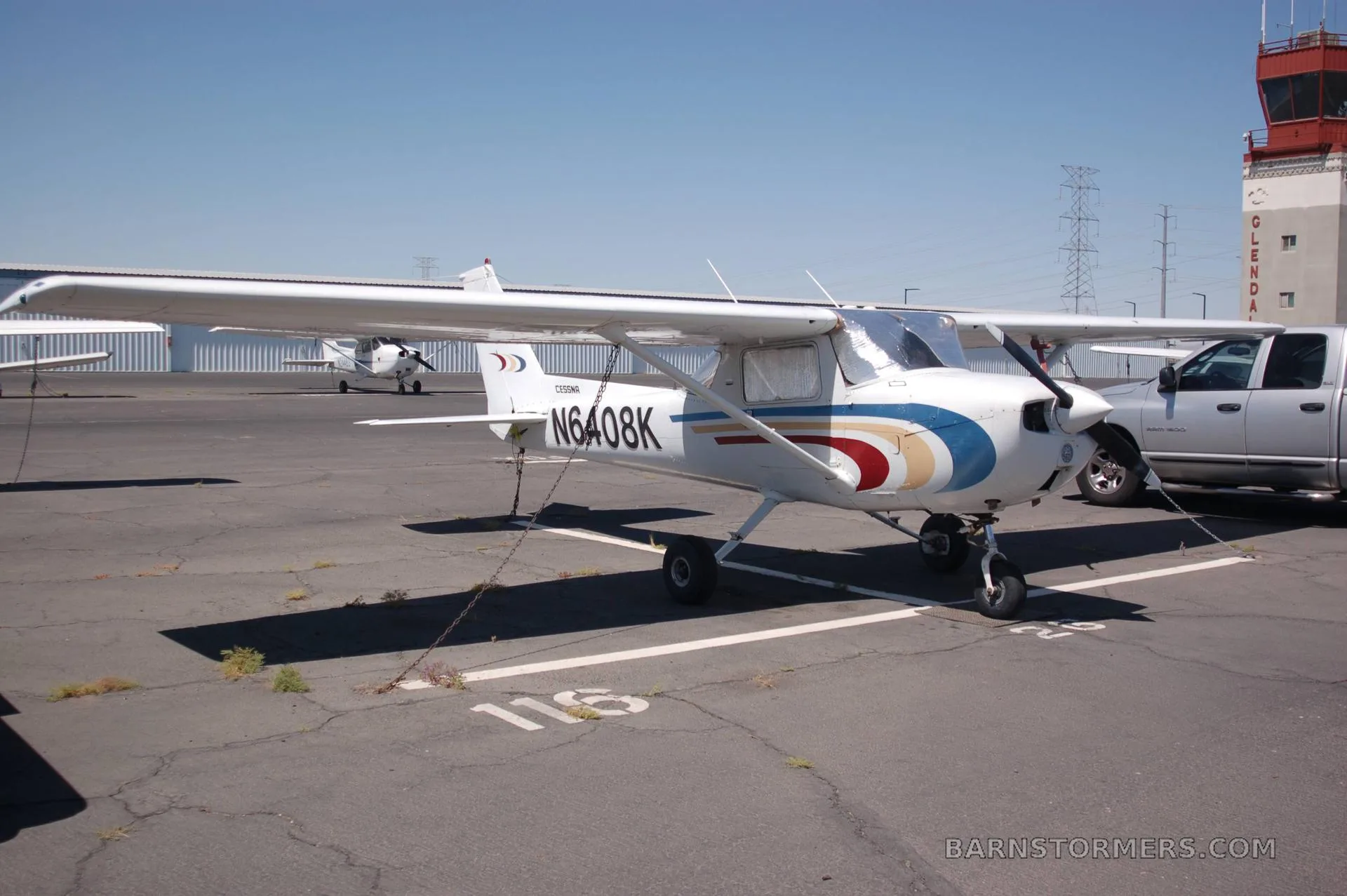 A white Cessna N6408K with blue, gold, and red stripes is tied down on an airfield next to a control tower.