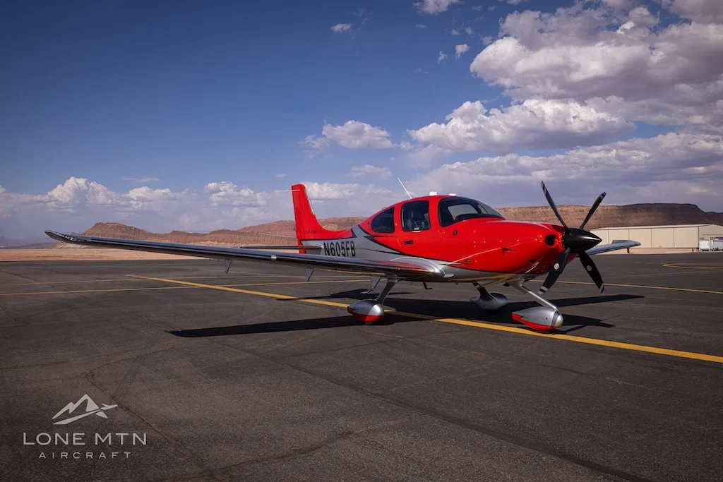 A front-quarter view of a red and silver Cirrus SR22T aircraft (N605FB) on an airfield with clouds in the sky.