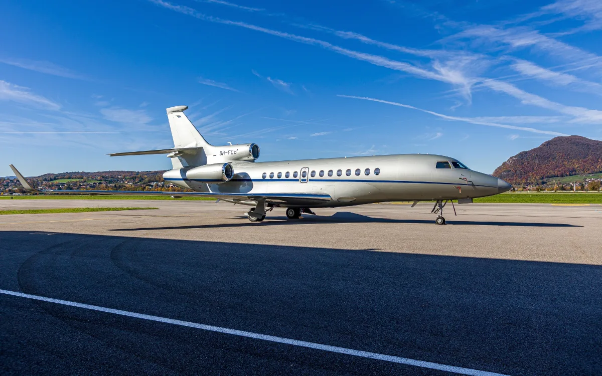 A silver Dassault Falcon 900 business jet with a blue stripe parked on an airport tarmac under a clear blue sky with mountains in the background.