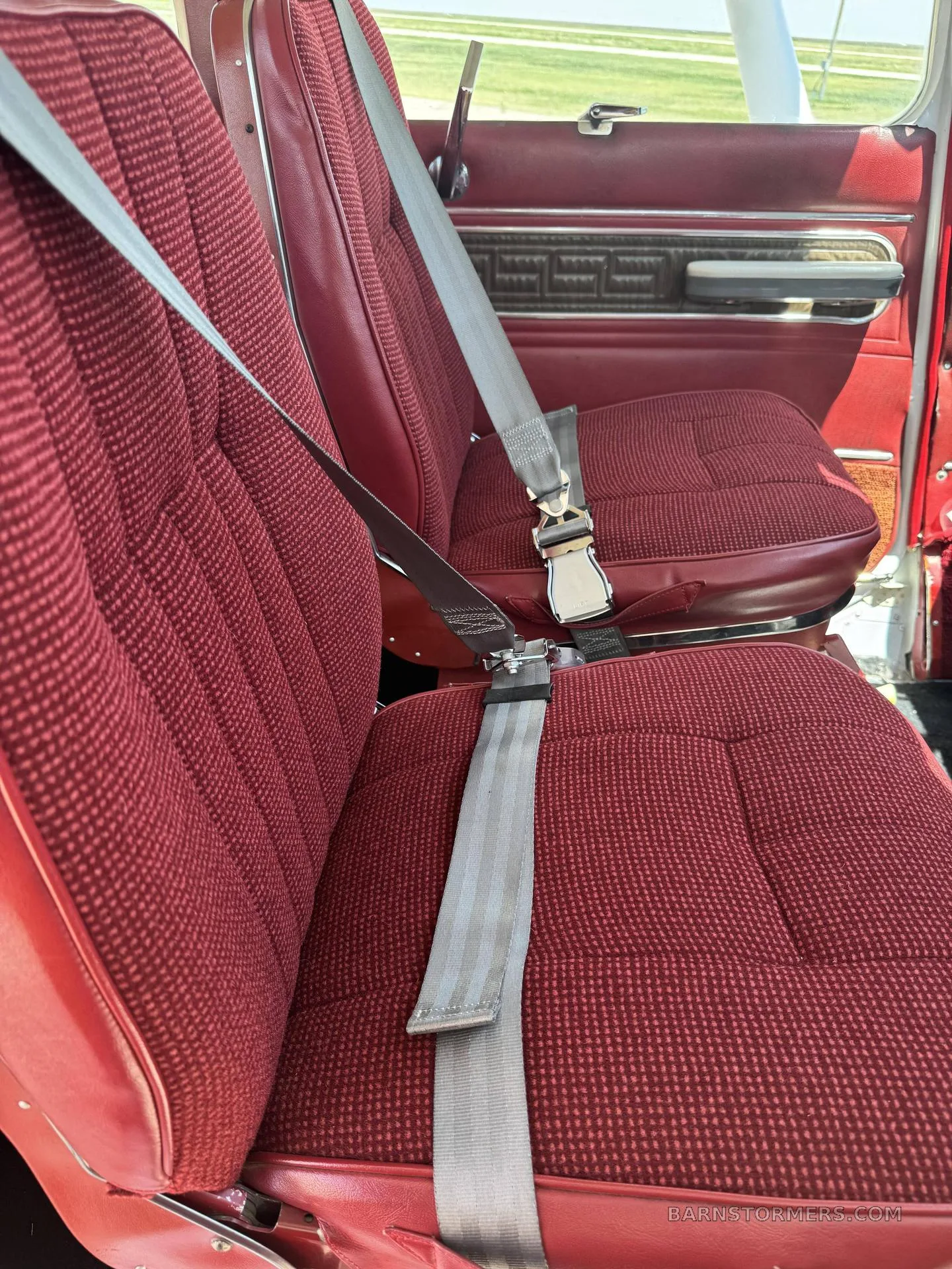 Two red fabric aircraft seats with grey seatbelts in the cabin, with a view towards the aircraft door.