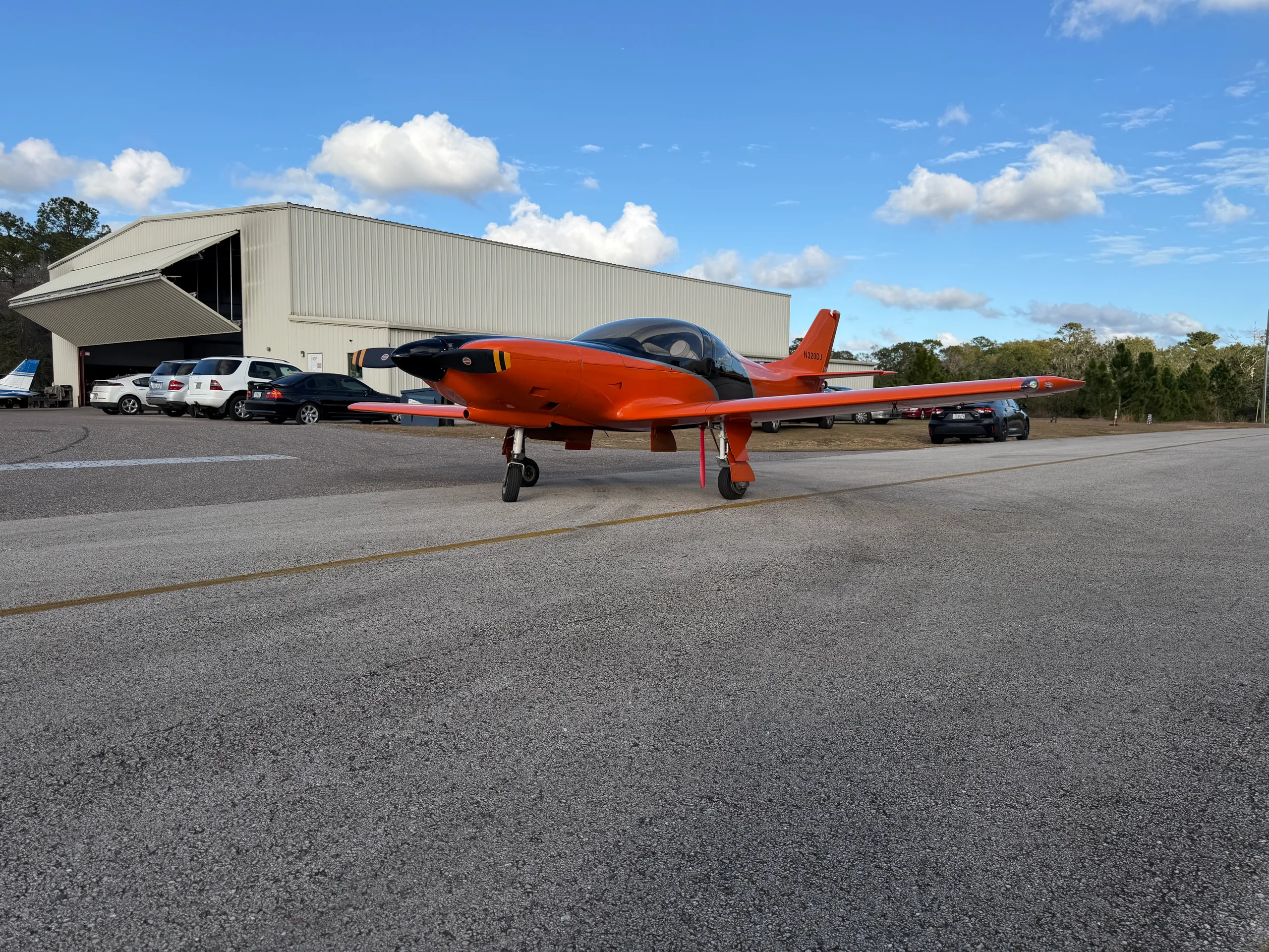 An orange and black experimental aircraft, N320DJ, sits on an airfield tarmac in front of a hangar under a partly cloudy sky.