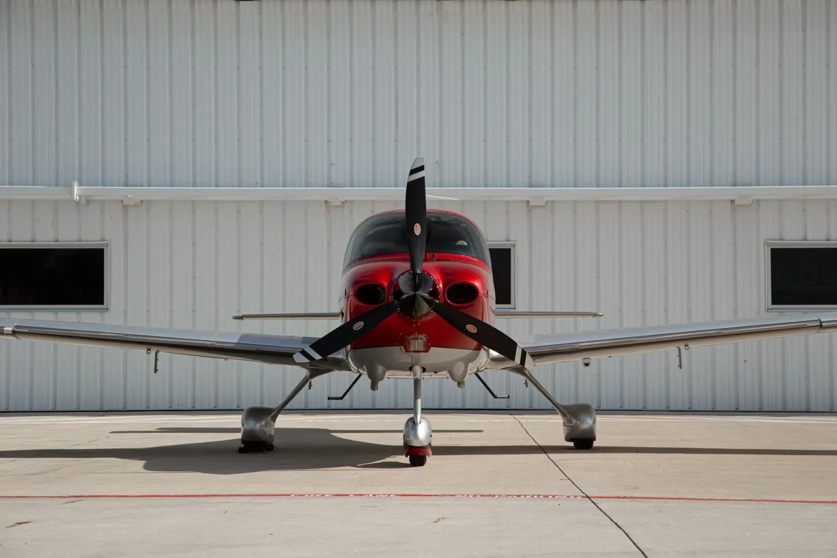 A head-on view of a red and silver single-engine propeller aircraft, N389WM, parked on an airfield in front of a white hangar.