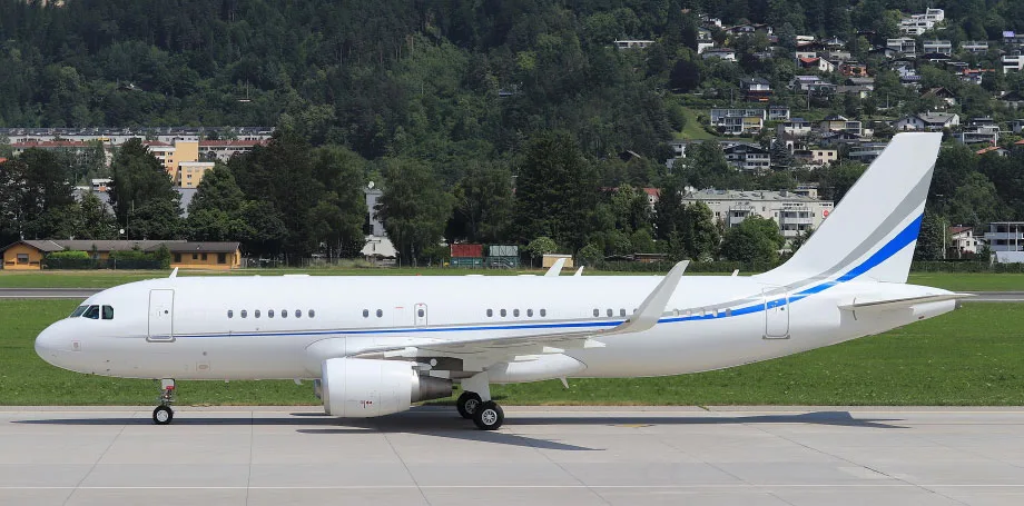 A white Airbus A319 with blue and grey stripes on the tail and fuselage is parked on an airport tarmac with mountains in the background.