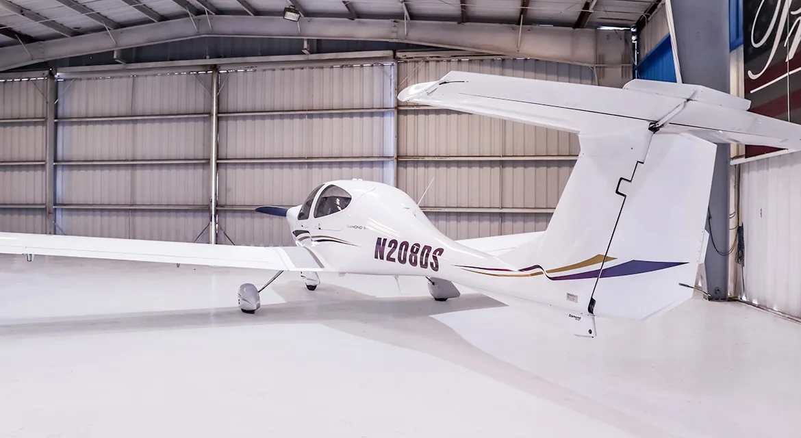 A rear-right view of a white Diamond DA40 Star aircraft, registration N208DS, with purple and gold tail stripes, in a hangar.