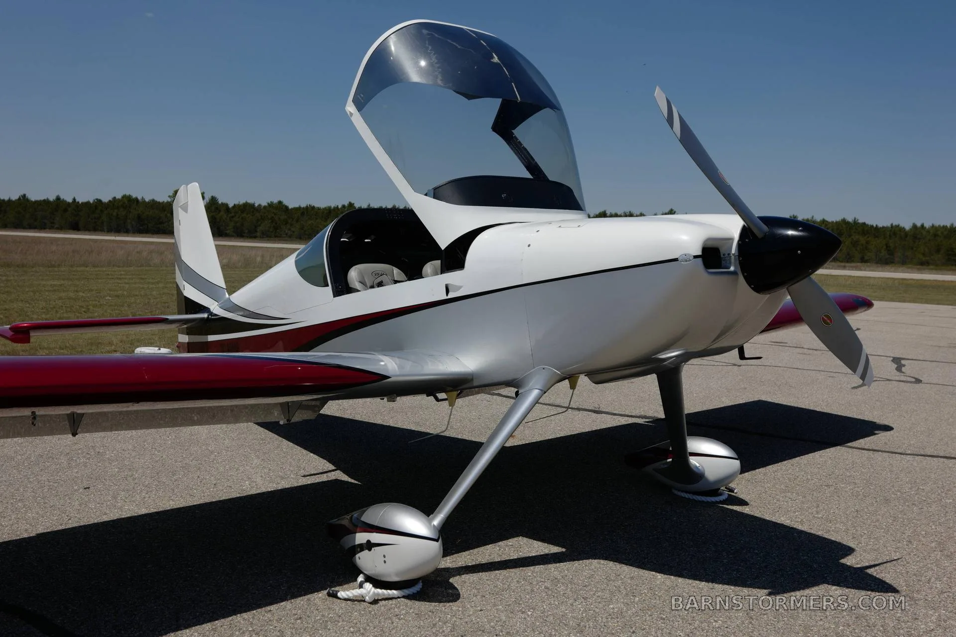 A white and red Vans RV-140MP aircraft with an open canopy, parked on an asphalt surface under a clear sky.