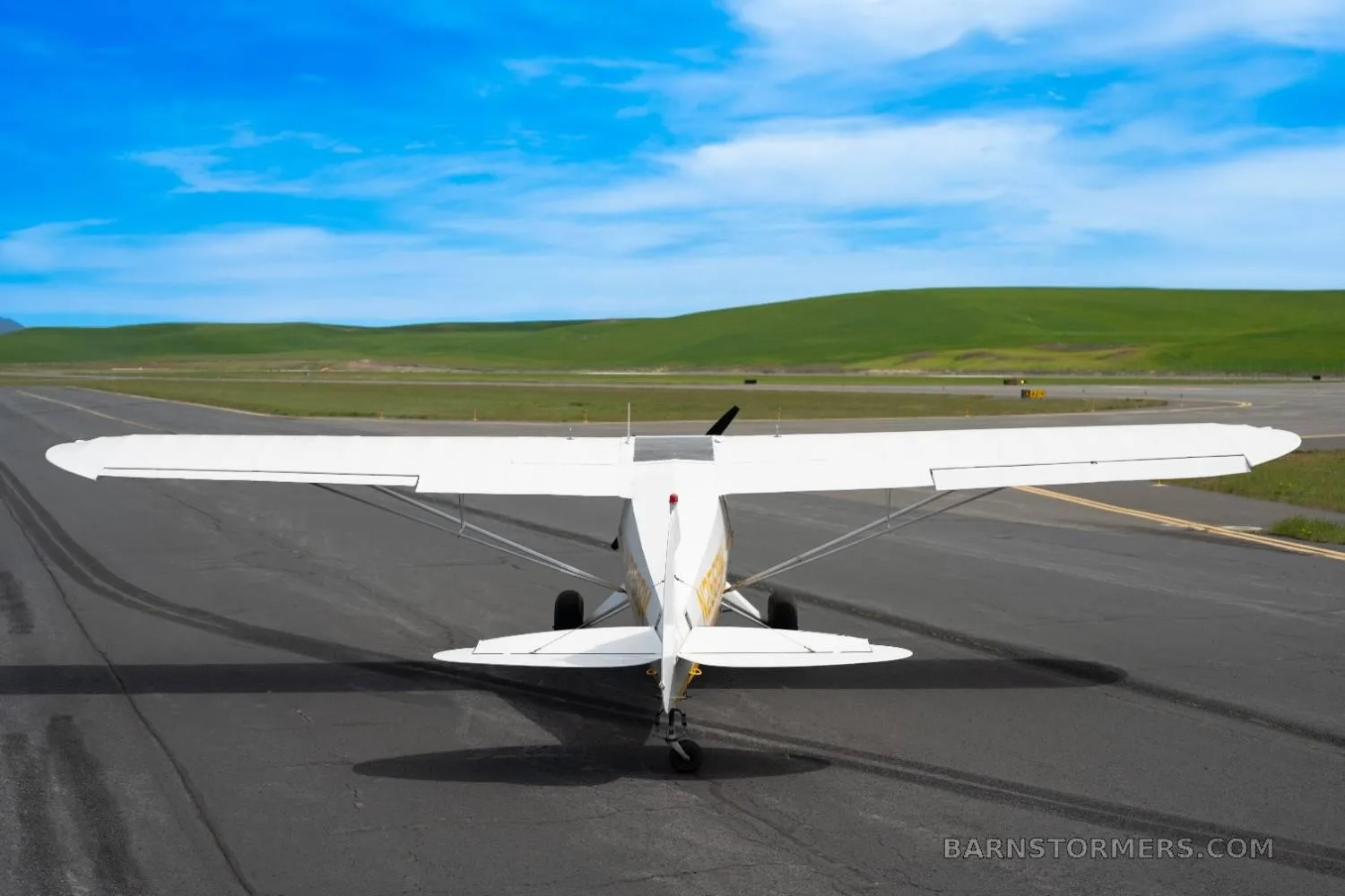 Rear view of a white and yellow single-engine taildragger aircraft (N2569M) on an asphalt runway under a blue sky.