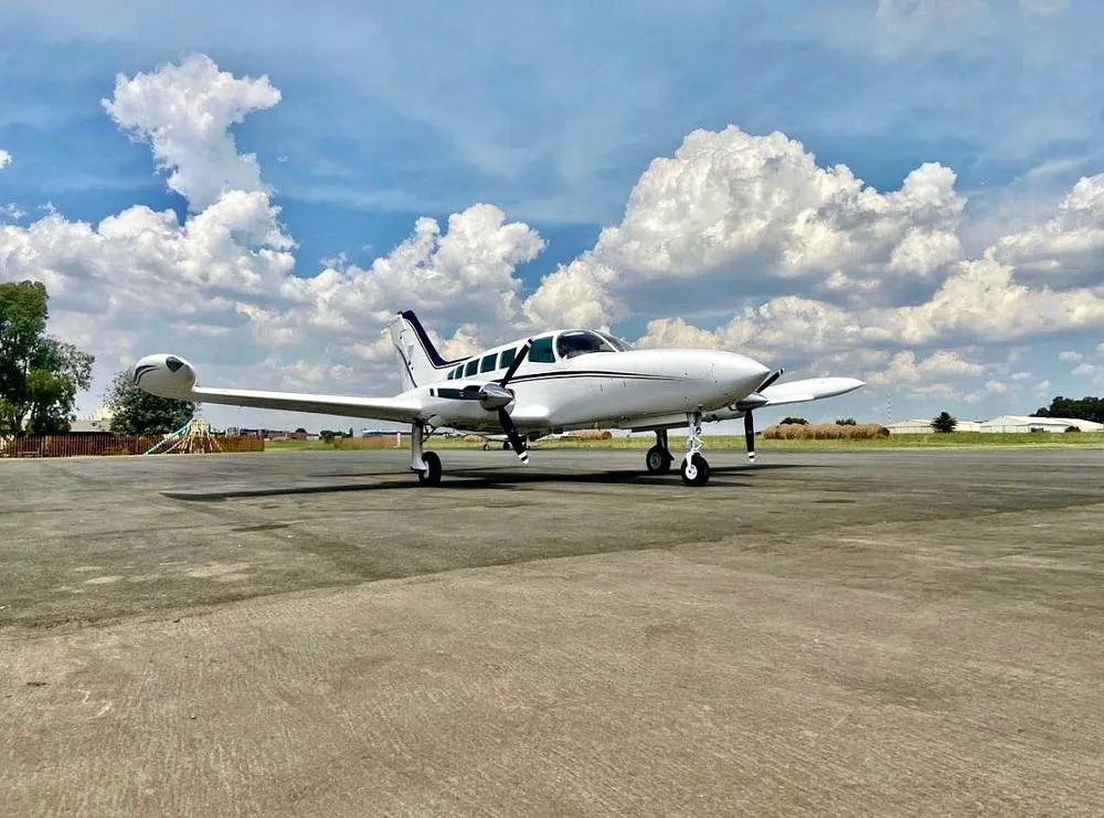 A white twin-engine propeller aircraft with dark blue stripes is parked on an asphalt tarmac under a partly cloudy sky.