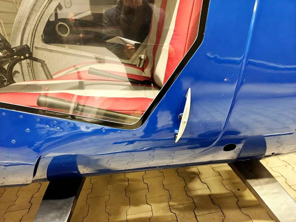 A close-up view of the blue fuselage and red and white striped seat of a helicopter, with a person reflected in the window.