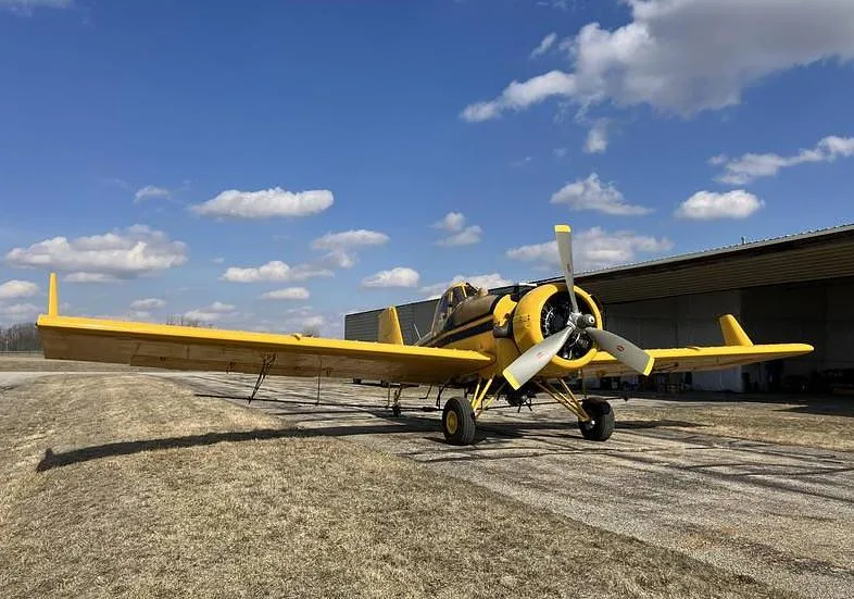 A yellow agricultural aircraft with black stripes is parked on an airfield next to a hangar under a partly cloudy sky.