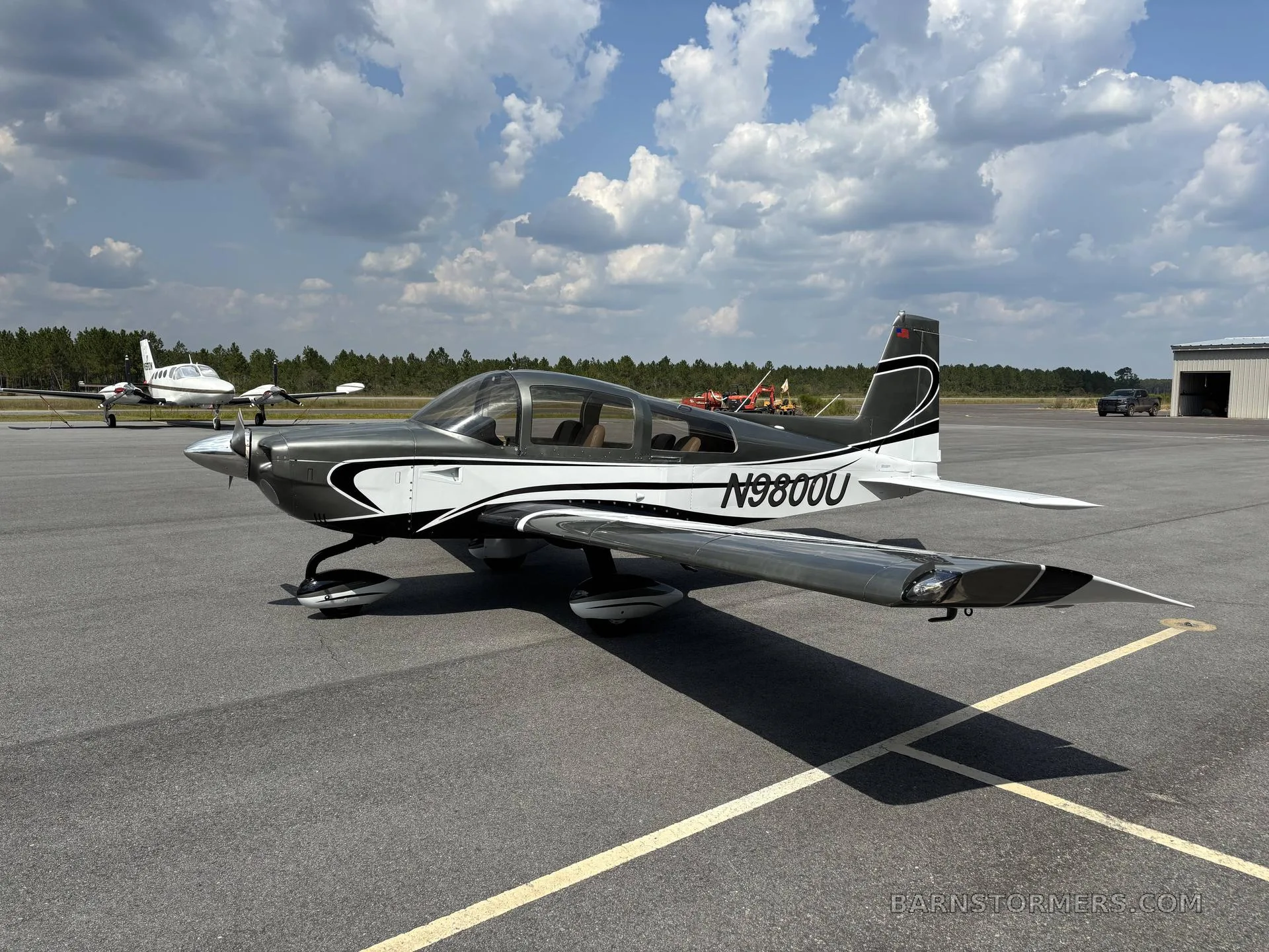 A gray and white Grumman American AA-5B Tiger (N9800U) parked on an asphalt ramp under a partly cloudy sky.
