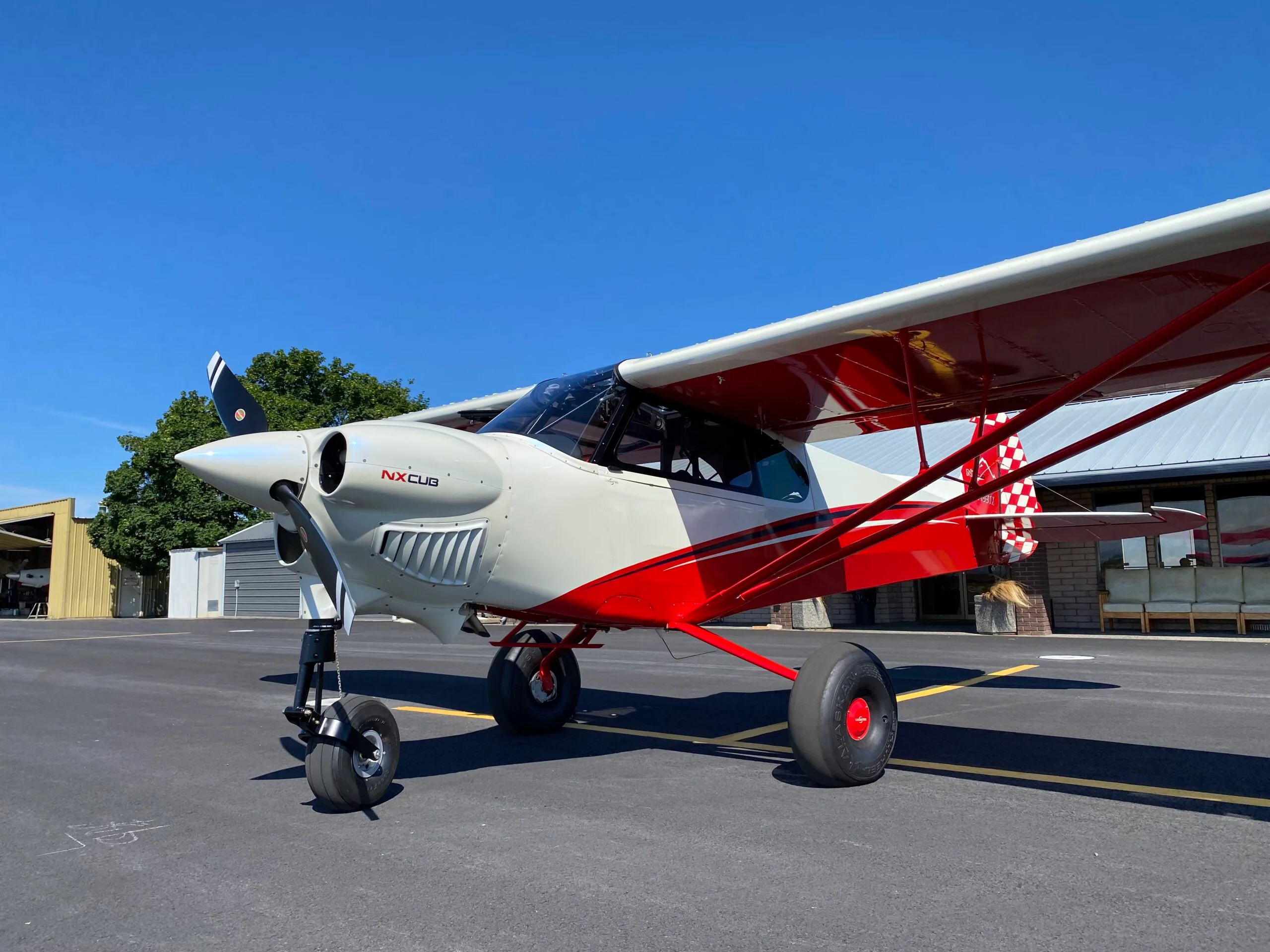 A white and red NX Cub aircraft with large tundra tires is parked on an asphalt tarmac under a clear blue sky, with hangars in the background.