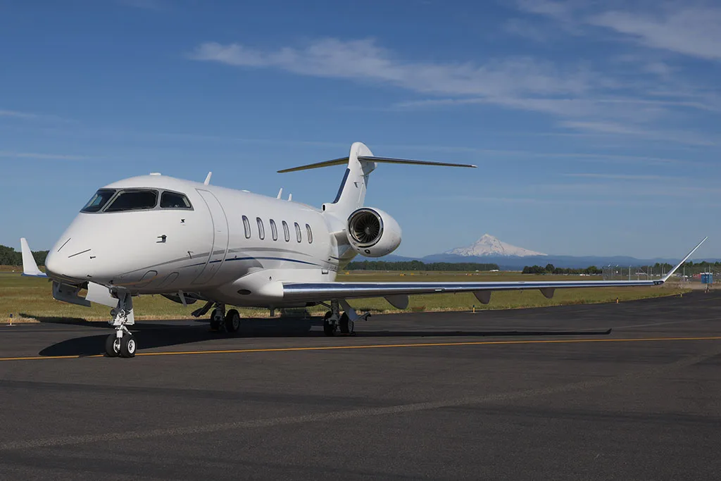 A white Bombardier Challenger 350 business jet parked on an airfield with a mountain in the distance under a clear blue sky.