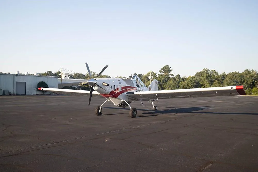 A white and red Thrush 510G aircraft parked on an asphalt tarmac with trees and buildings in the background.
