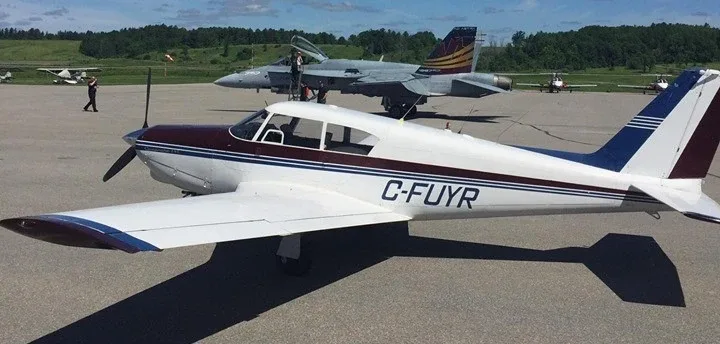 A white single-engine propeller aircraft with maroon and blue stripes, registration C-FUYR, parked on an airport tarmac with other planes in the background.