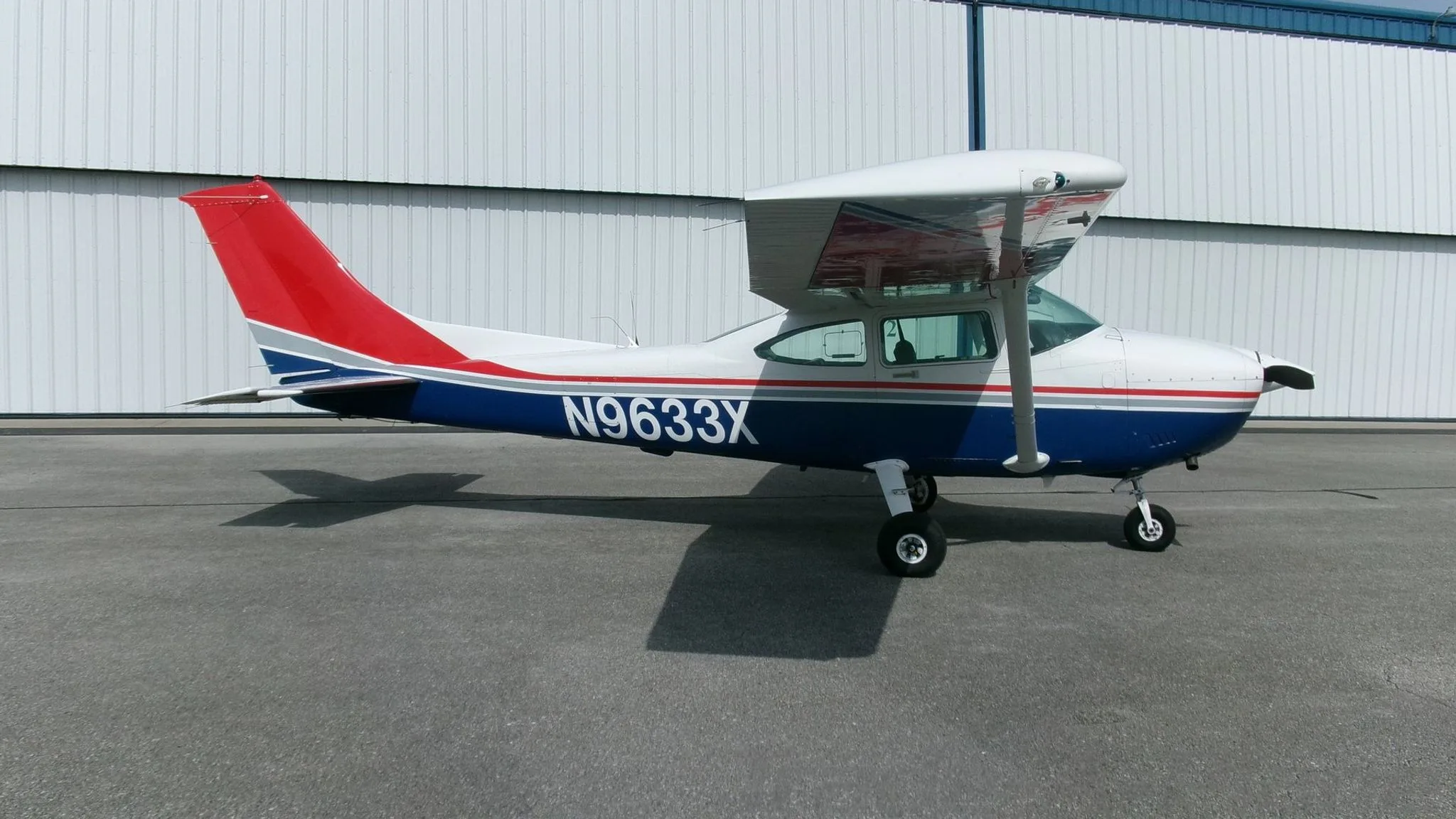 A side view of a white, blue, and red Cessna 182 Skylane with registration N9633X parked on an asphalt apron.
