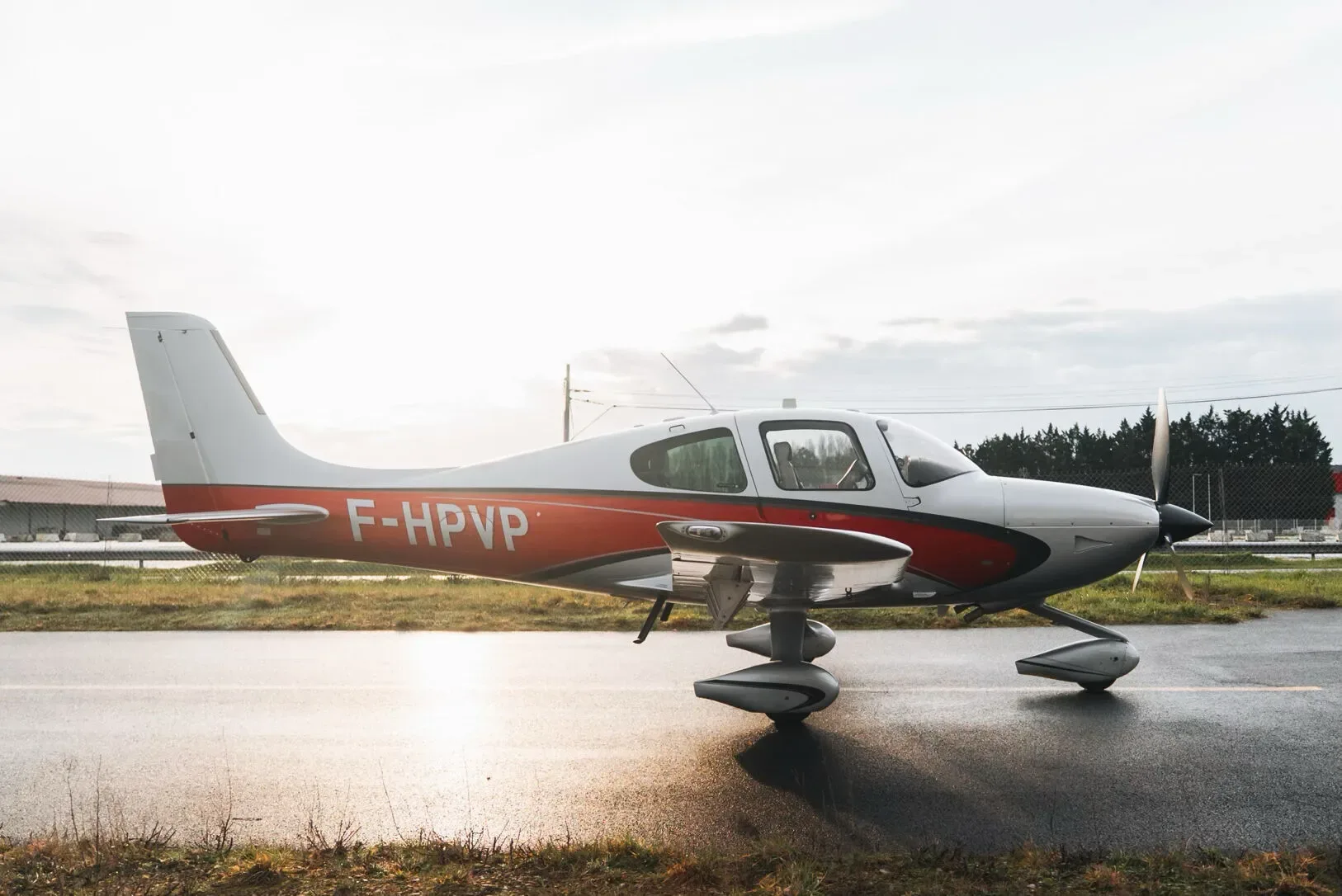 A silver and red Cirrus SR22 aircraft parked on an airfield with the sun low in the sky.