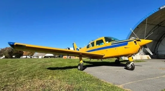 A yellow and blue single-engine aircraft parked on grass next to an asphalt path and a hangar under a clear blue sky.
