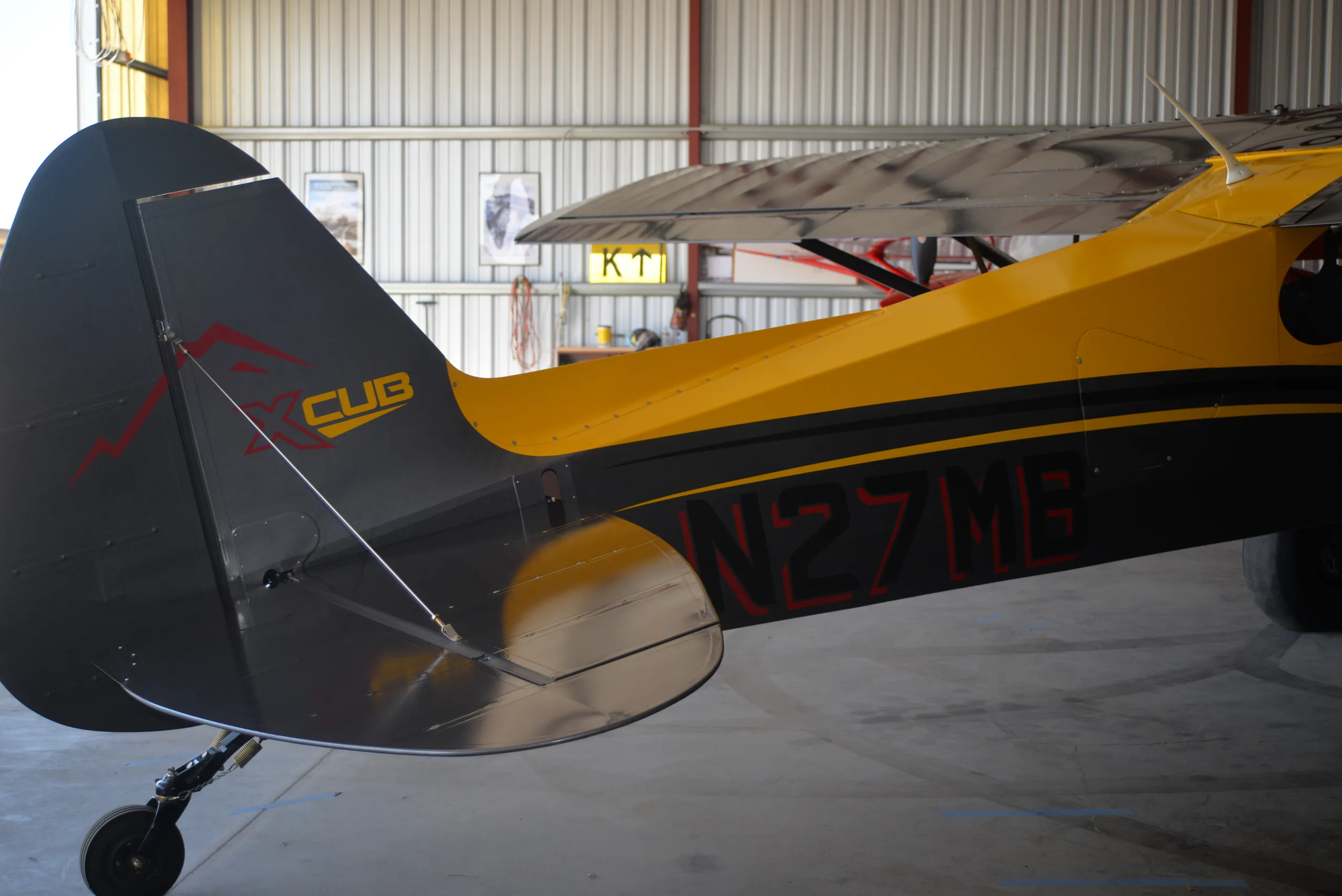 The tail of a yellow and gray CubCrafters XCub, registration N27MB, inside a hangar.