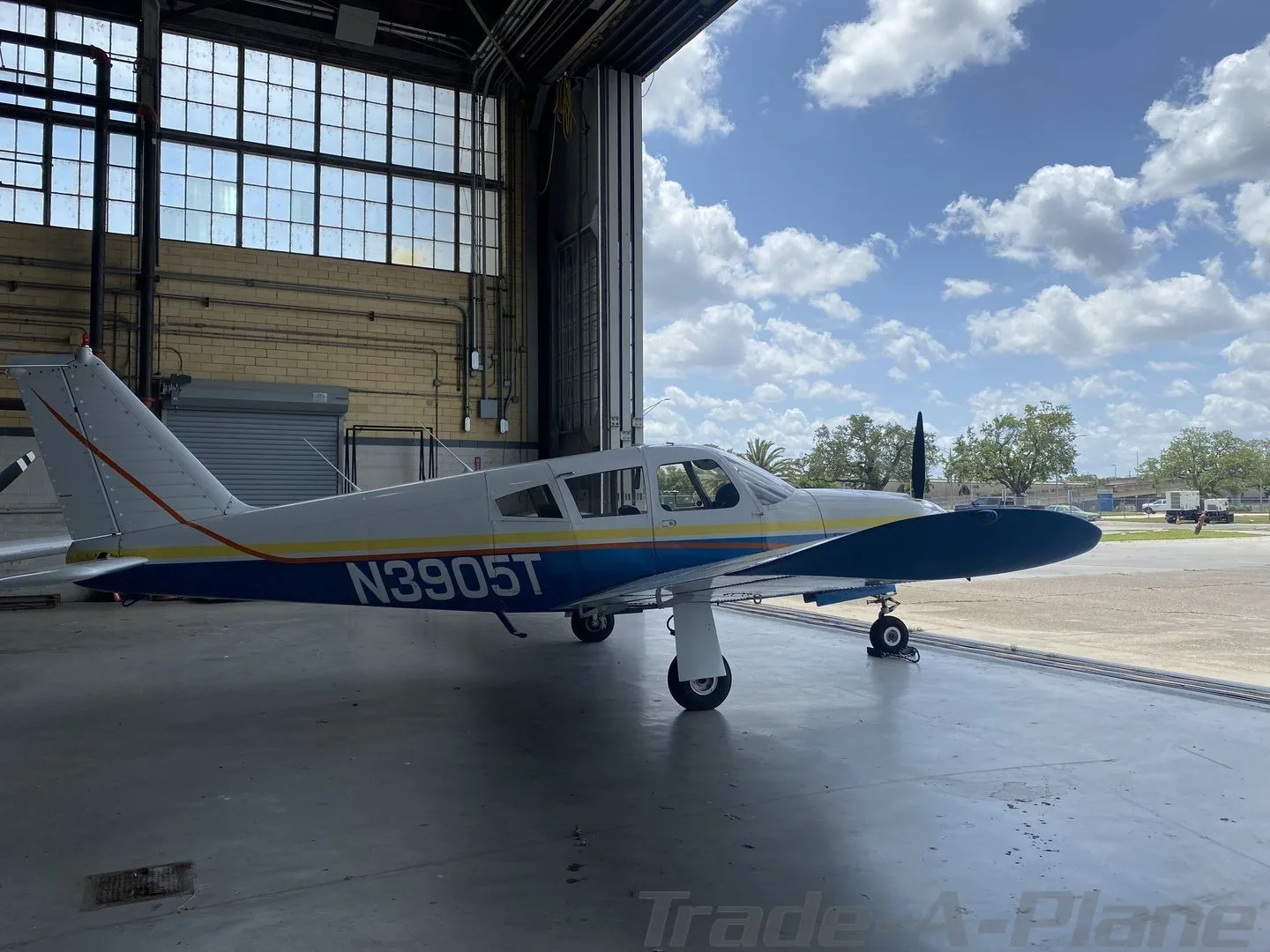 A Piper Cherokee-style aircraft, N3905T, with a white, blue, and yellow paint scheme, parked partially inside a hangar on a sunny day.