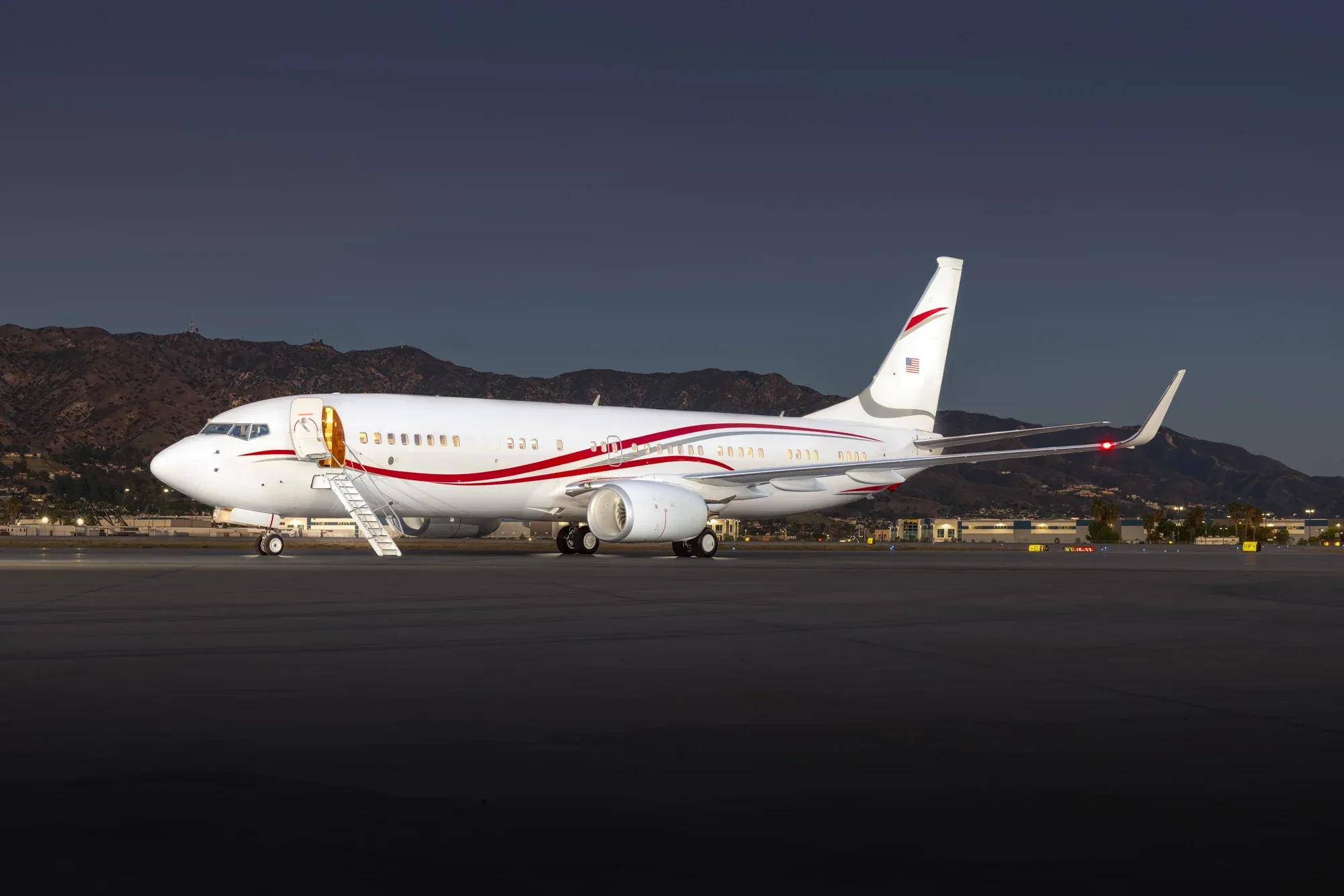 A white Boeing Business Jet with red and grey stripes is parked on a tarmac at dusk, with mountains in the background.