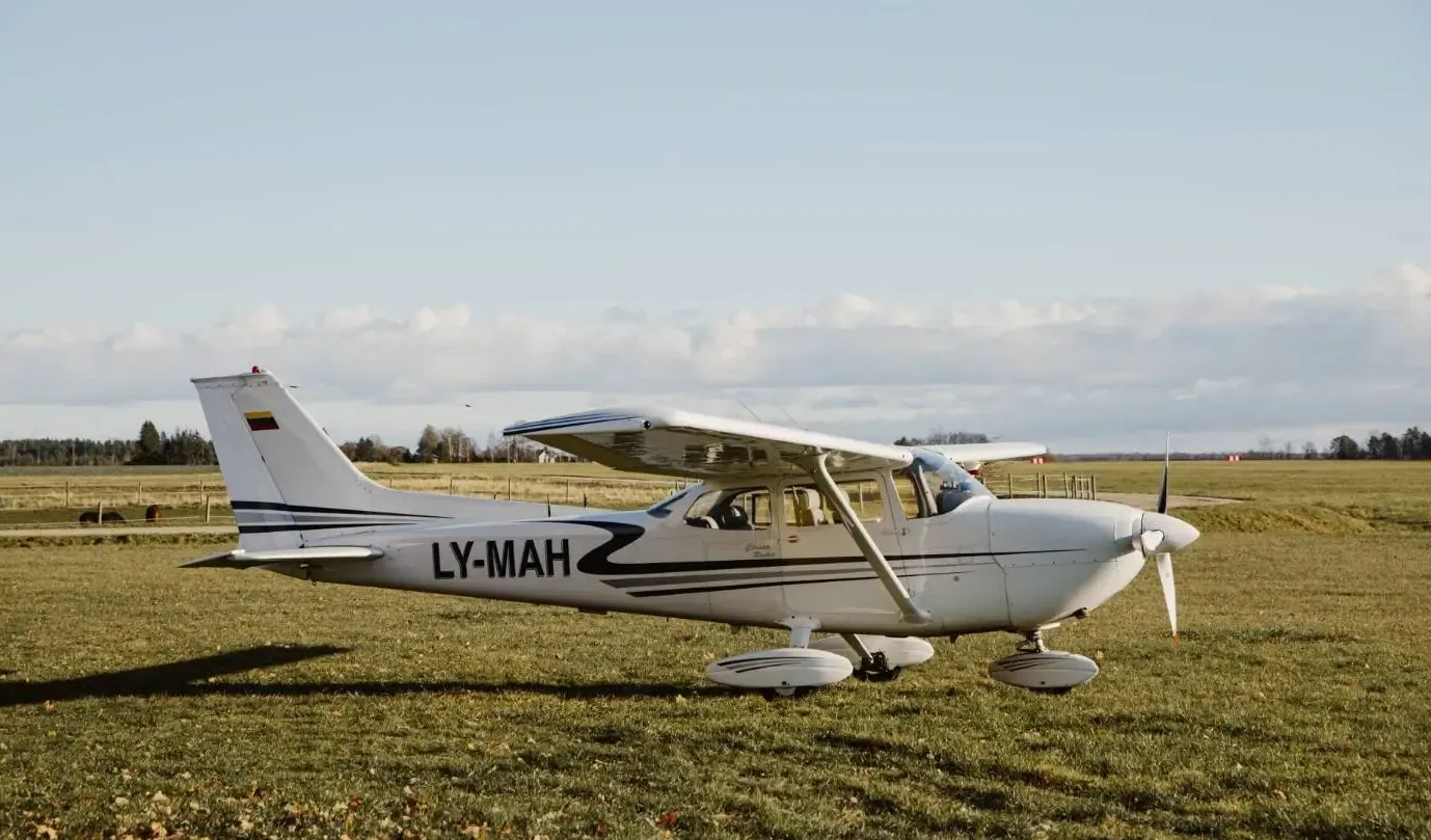 A white Cessna 172 with black and grey stripes, registration LY-MAH, parked on a grassy field with trees in the background.