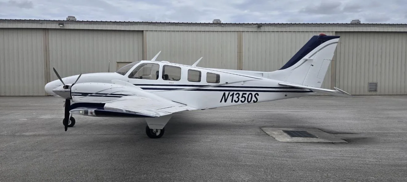 A white Beechcraft Baron N1350S with blue and grey stripes parked on a tarmac in front of a hangar.