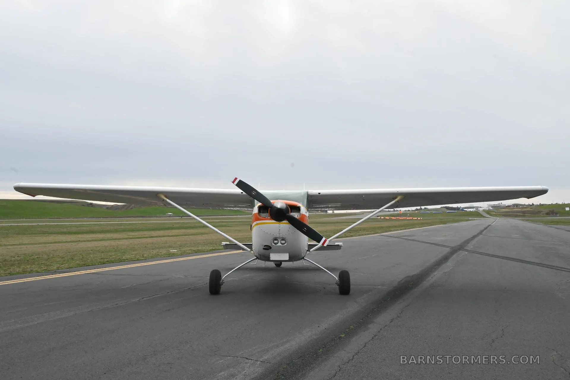 A front view of a white and orange Cessna 172 Hawk XP II (N5205V) on an asphalt runway under a cloudy sky.