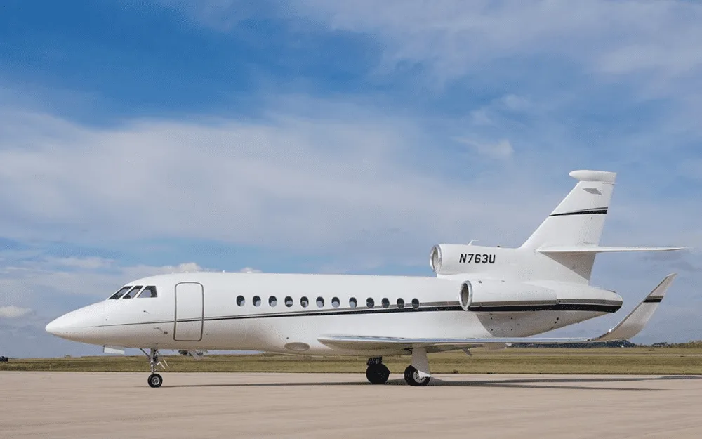 A white Dassault Falcon 900EX business jet with black and grey stripes on the tail, parked on an airfield under a blue sky.