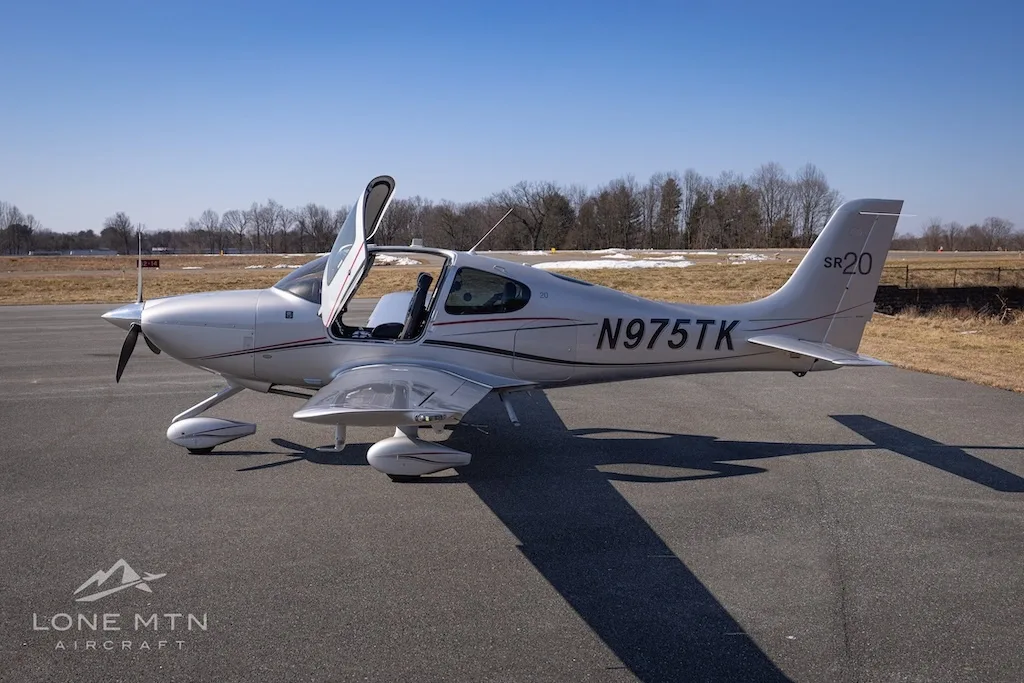 A silver Cirrus SR20 aircraft with registration N975TK parked on an asphalt runway, viewed from the left side with doors open.