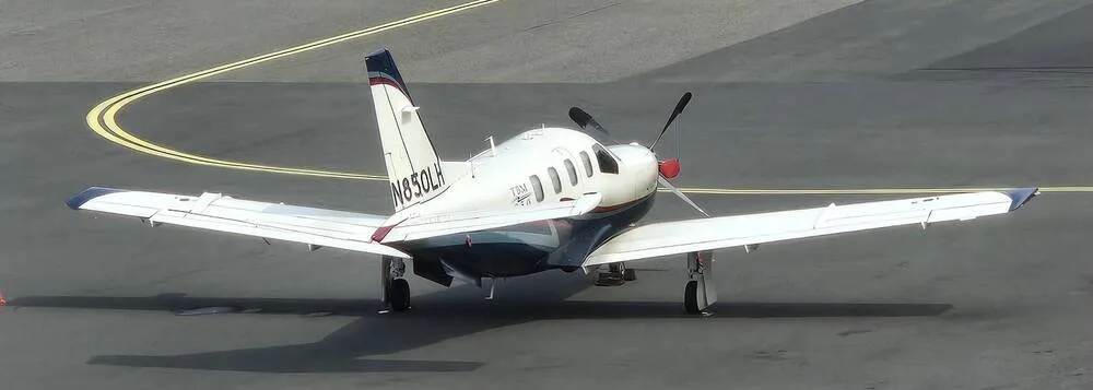 An aerial view of the white TBM 850 aircraft with blue and red stripes, parked on an asphalt tarmac.