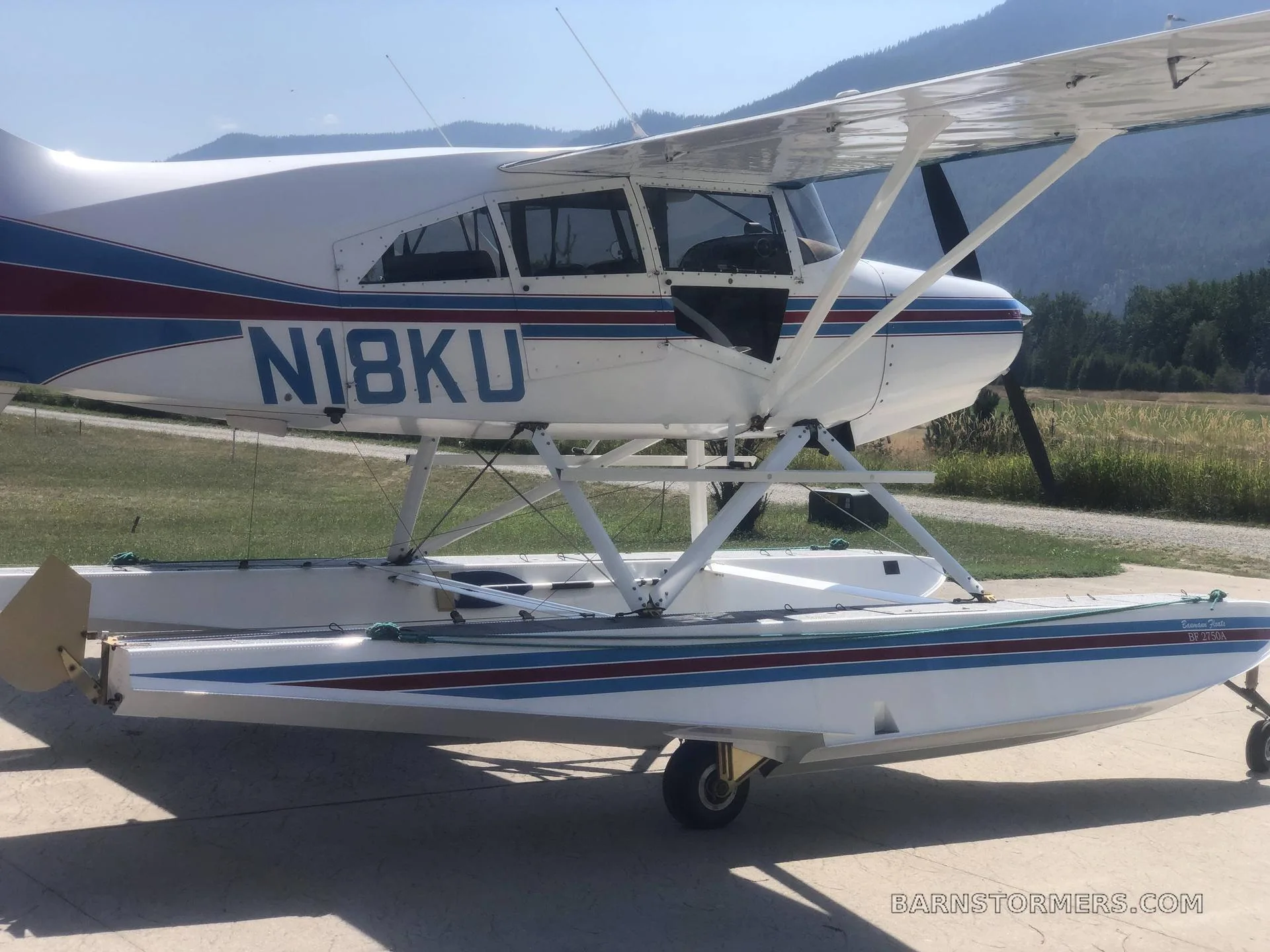 A white and blue floatplane, N18KU, with red tail accents, parked on concrete with mountains in the background.