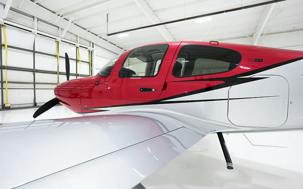 A close-up view of the red and white Cirrus SR22 GTS aircraft's fuselage and wing inside a hangar.