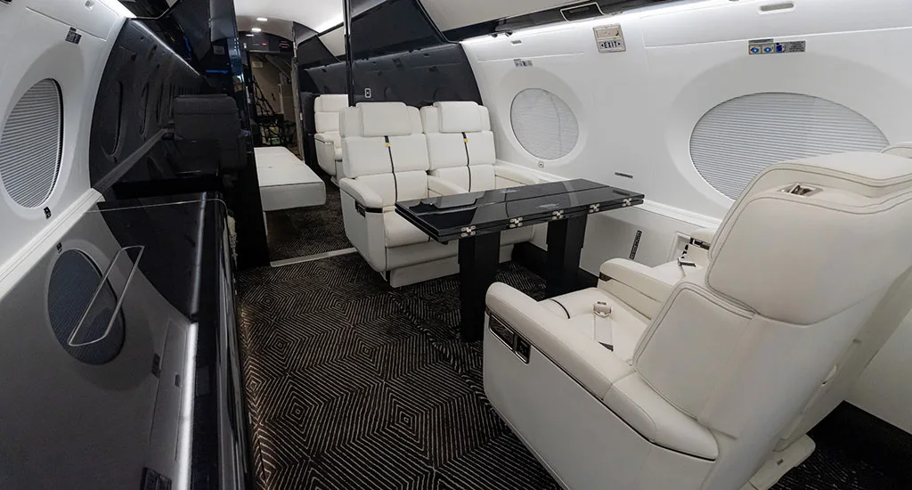 A view inside the business jet cabin showing white leather seats, a black table, and dark patterned carpet.