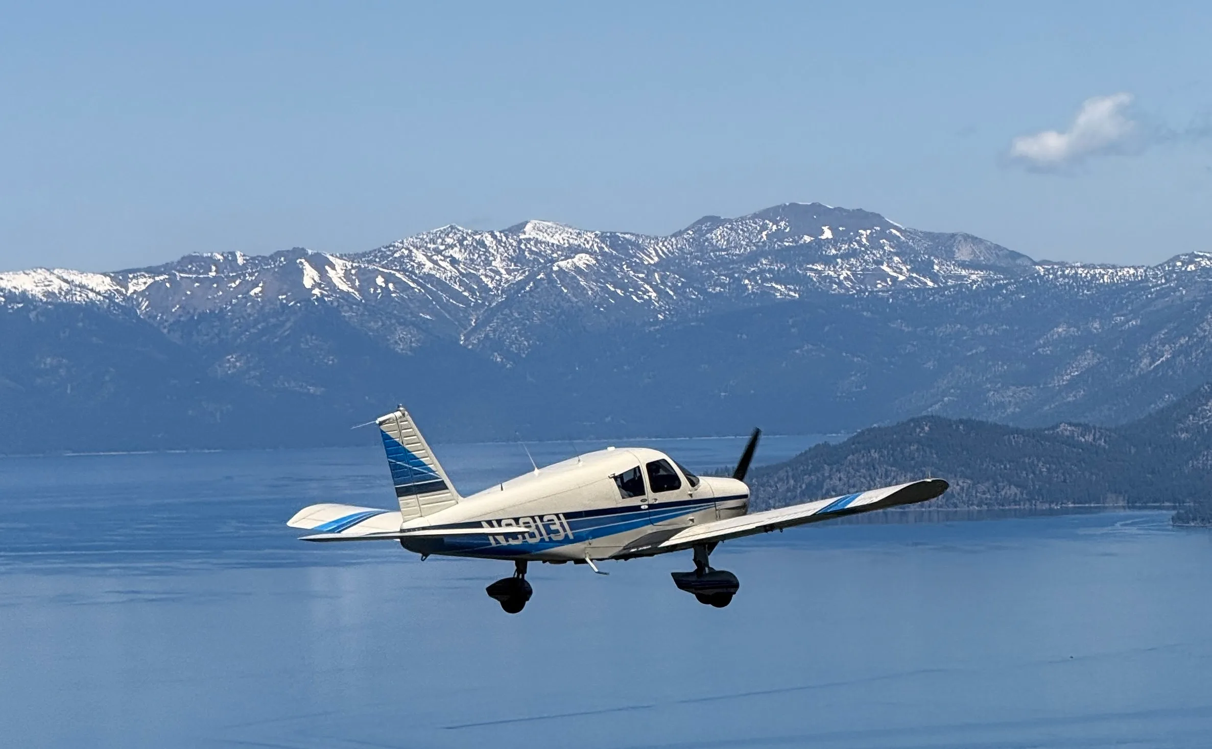 A white and blue Piper Cherokee aircraft (N98131) flies over a lake with snow-capped mountains in the background.