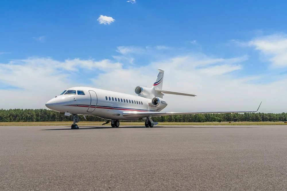 A front-side view of a white Dassault Falcon 900EX business jet with red and blue stripes, parked on an airfield under a blue sky.
