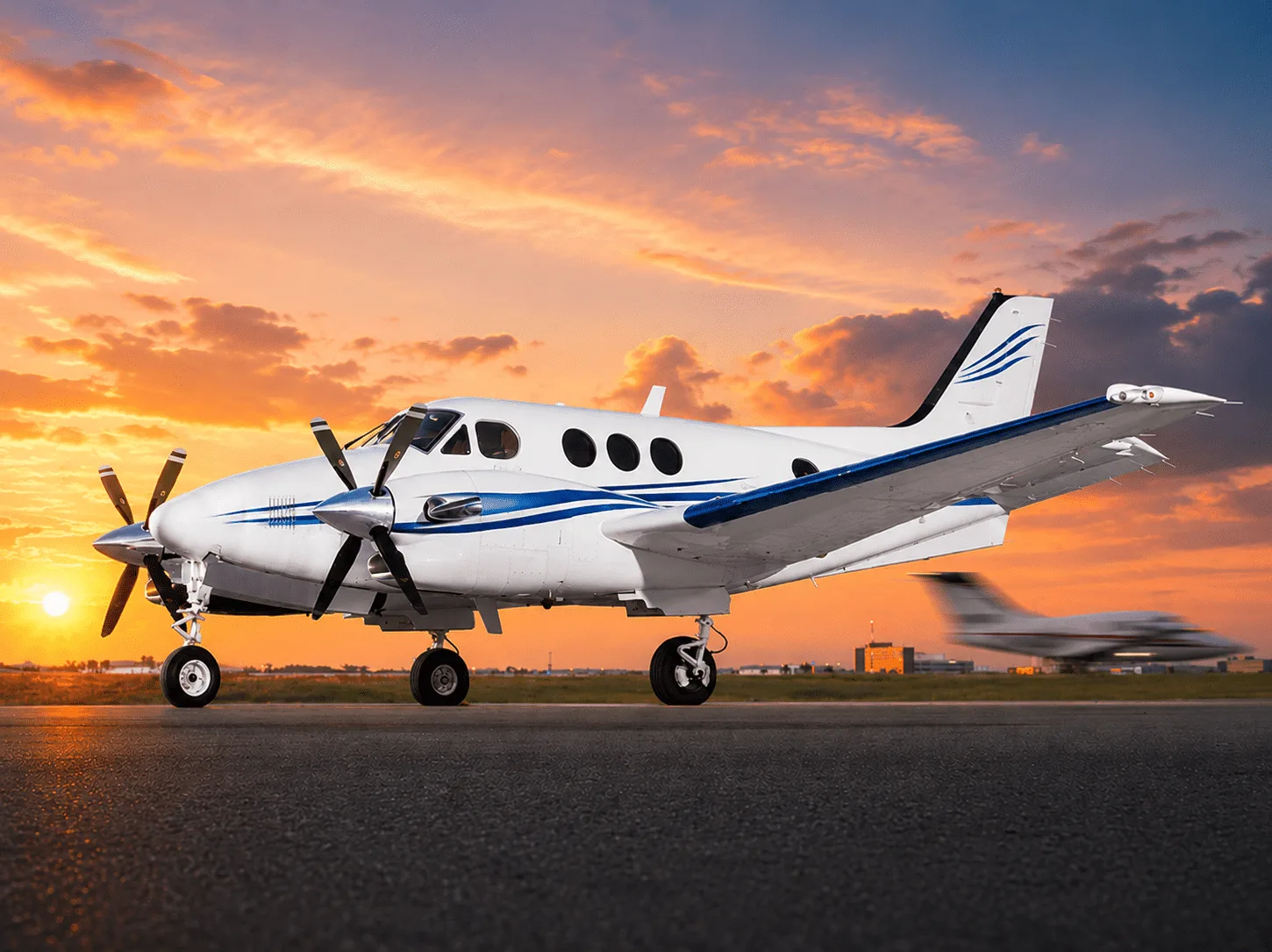 A white twin-turboprop aircraft with blue stripes is parked on a runway at sunset, with another aircraft blurred in the background.