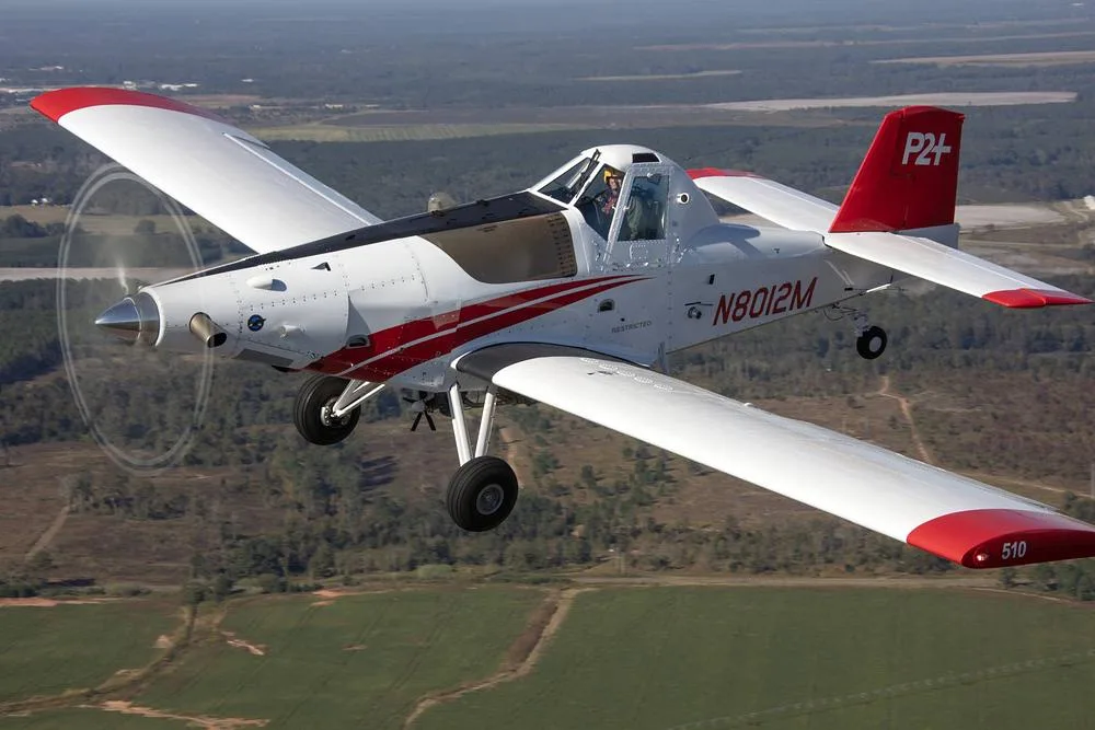 An aerial view of a white and red crop duster aircraft (N8012M) in flight over a rural landscape.