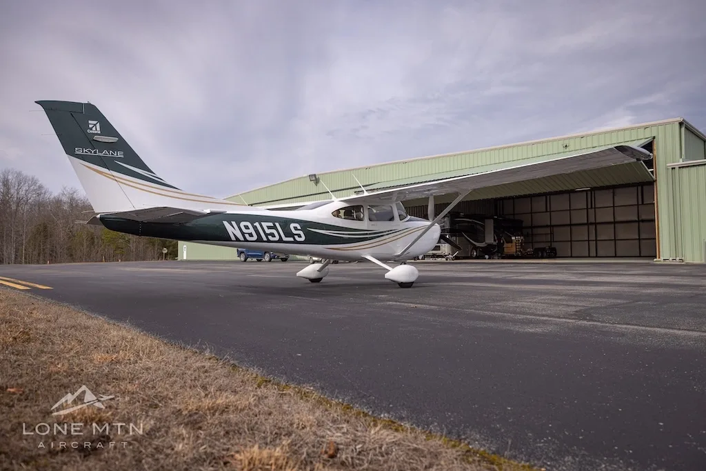 A white and dark green Cessna Skylane N915LS with gold stripes, parked on an asphalt ramp in front of an open hangar.