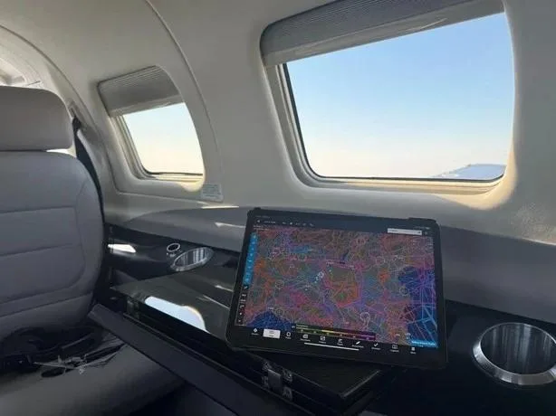The interior cabin of an aircraft with light-colored seats, windows showing the sky, and a tablet displaying a map on a dark table.