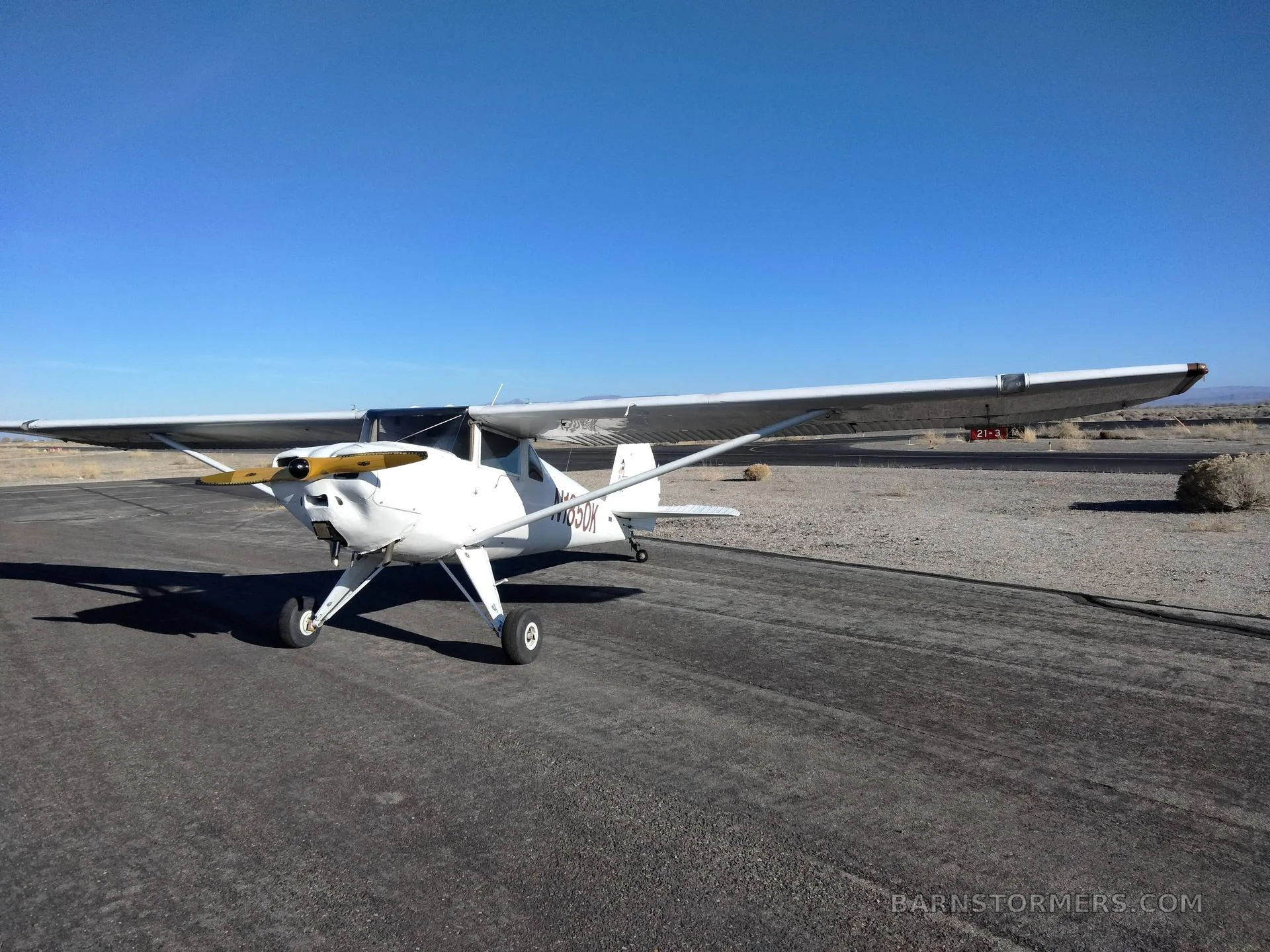 A white Luscombe 8A aircraft with a yellow propeller is parked on an asphalt runway in a wide shot under a clear blue sky.