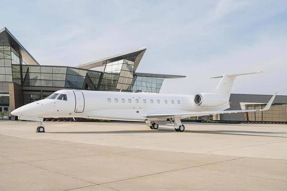 A white Embraer Legacy business jet is parked on an airport tarmac in front of a modern glass building.