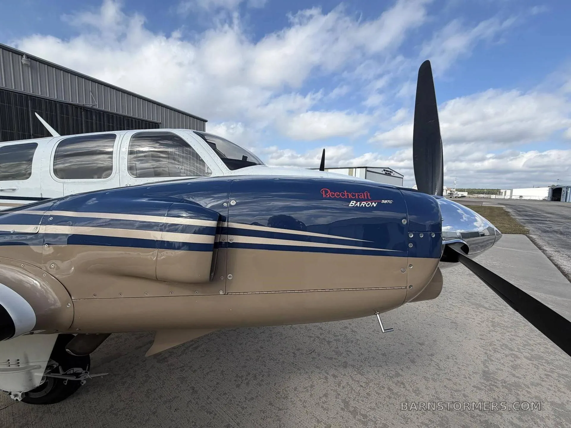 A close-up of the blue and tan nose and propeller of a Beechcraft Baron 58TC aircraft, parked on an airfield next to a hangar.