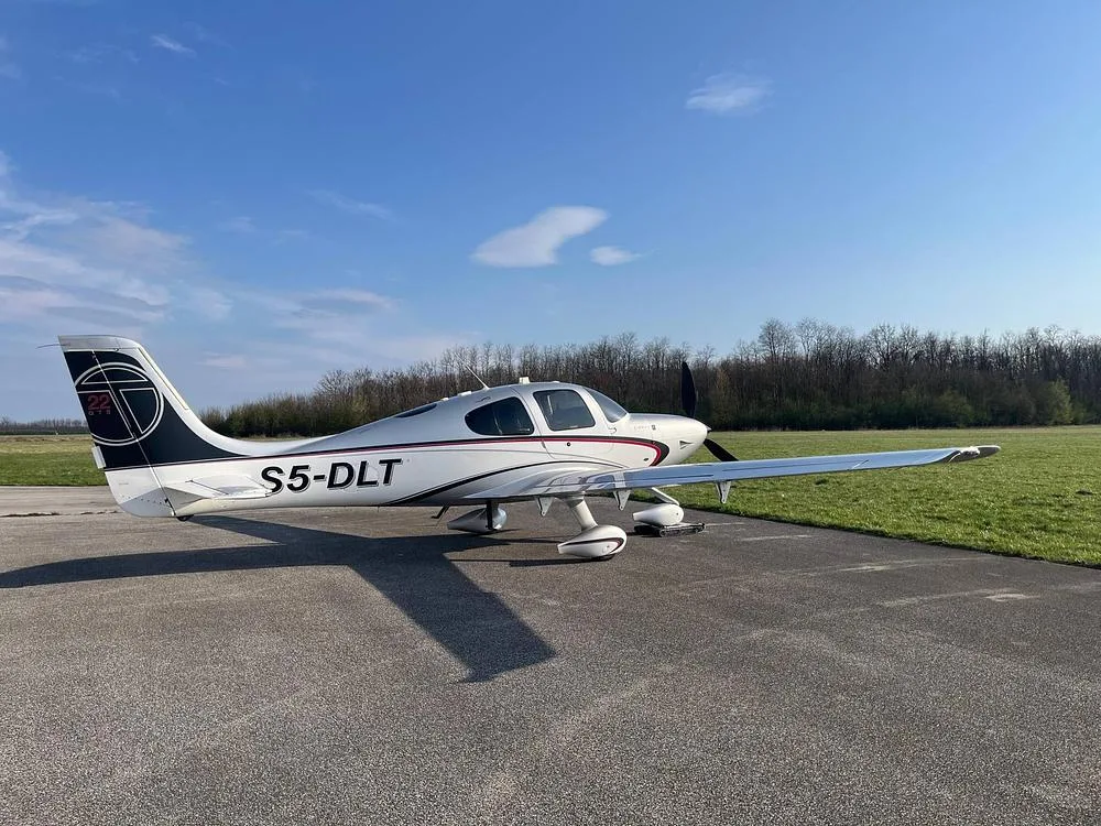 A silver and white Cirrus SR22T aircraft with registration S5-DLT parked on an asphalt surface next to a grassy field.