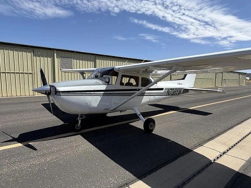 A front-left view of the white Cessna 172, N738YW, with gray and black stripes, parked on a tarmac next to a hangar.